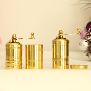 May include: Two gold metal pepper mills with a crank handle. The mills are separated and the top portion of one mill is removed, revealing the grinding mechanism.