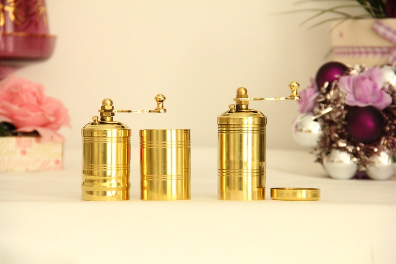 May include: Two gold metal pepper mills with a crank handle. The mills are separated and the top portion of one mill is removed, revealing the grinding mechanism.