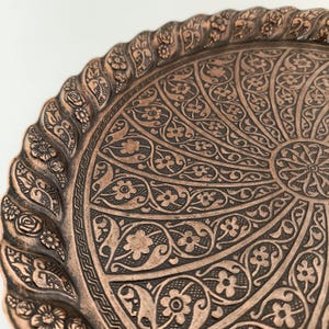May include: A round copper tray with an intricate floral design. The tray has a raised edge with a decorative border.