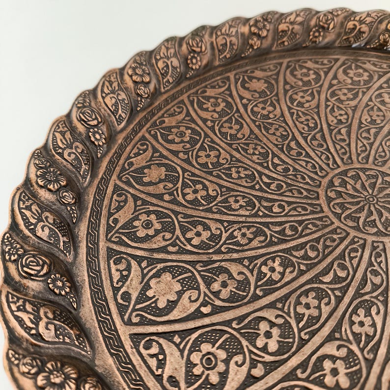 May include: A round copper tray with an intricate floral design. The tray has a raised edge with a decorative border.