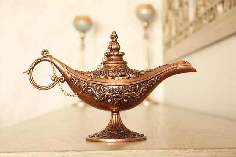 Peut inclure: Une lampe &agrave; genie de couleur dor&eacute;e avec des motifs floraux complexes et une poign&eacute;e en cha&icirc;ne. La lampe est sur une surface en bois clair.