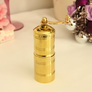 May include: A gold metal pepper grinder with a crank handle. The grinder has a cylindrical shape and is made of polished metal.