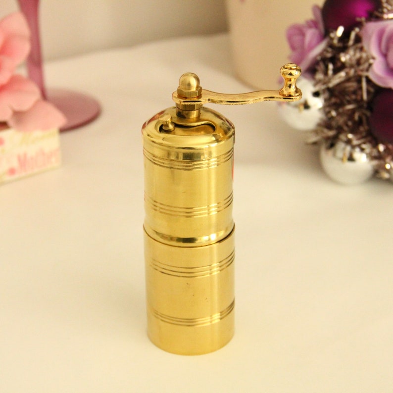 May include: A gold metal pepper grinder with a crank handle. The grinder has a cylindrical shape and is made of polished metal.