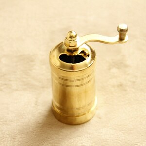 May include: A gold metal pepper grinder with a crank handle. The grinder has a cylindrical shape and a smooth, polished finish.