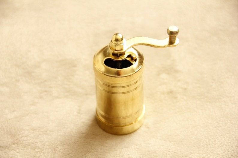 May include: A gold metal pepper grinder with a crank handle. The grinder has a cylindrical shape and a smooth, polished finish.