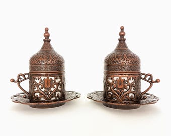 May include: Two ornate copper Turkish coffee cups with lids and saucers. The cups and saucers have intricate floral designs and are decorated with a white enamel.