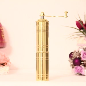 May include: A gold-colored pepper grinder with a hand crank, standing upright. The grinder has a cylindrical body with horizontal etched lines. A decorative arrangement of flowers and ornaments is in the background.