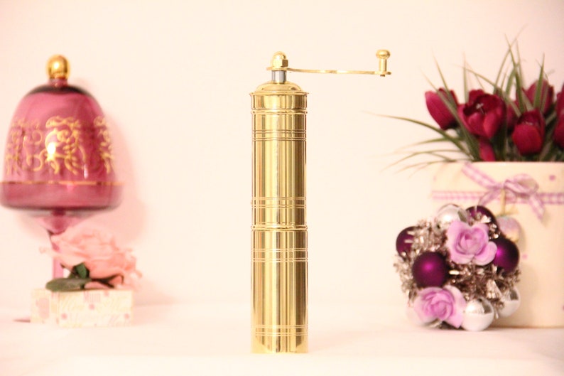 May include: A gold-colored pepper grinder with a hand crank, standing upright. The grinder has a cylindrical body with horizontal etched lines. A decorative arrangement of flowers and ornaments is in the background.