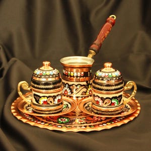 Pode incluir: Um conjunto de café de cobre com duas chávenas pequenas, uma cafeteira e uma bandeja. As chávenas e a cafeteira são adornadas com desenhos florais pretos, dourados e coloridos. O conjunto tem uma pega de madeira castanha escura e assenta num fundo escuro.