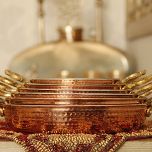 May include: A set of six copper serving dishes with hammered texture and gold handles. The dishes are stacked on top of each other, with the largest dish at the bottom and the smallest dish at the top.