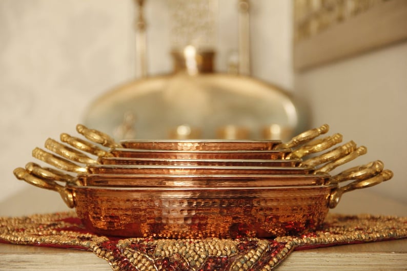 May include: A set of six copper serving dishes with hammered texture and gold handles. The dishes are stacked on top of each other, with the largest dish at the bottom and the smallest dish at the top.
