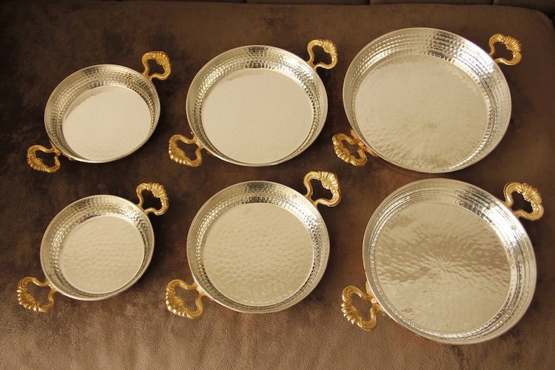 May include: Set of six silver-colored metal serving dishes with gold-colored handles. The dishes are round and have a hammered texture.