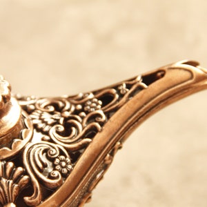 Peut inclure: Une lampe &agrave; genie de couleur dor&eacute;e avec des motifs floraux complexes. La lampe a un sommet rond avec une petite boucle sur le dessus.