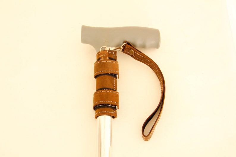 May include: A brown leather cane strap with a silver clasp and a gray handle. The strap is wrapped around the cane and has a loop for carrying the cane. Walking stick. Walking Stick Wrist Strap. Walking Cane. Cane Wrist Strap