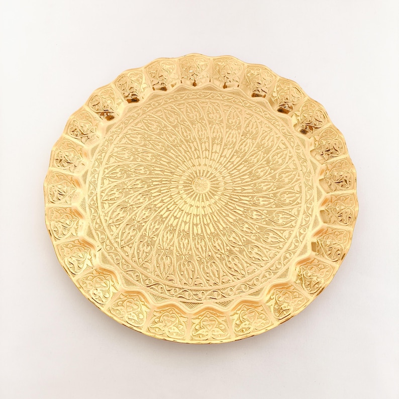 May include: A round, gold-colored metal serving tray with an intricate, embossed design. The tray has a scalloped edge and a raised center.