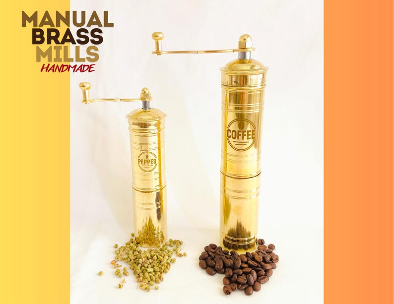 May include: Two brass manual mills, one labeled "Pepper" and the other "Coffee", with a crank handle. The mills are on a white surface with peppercorns and coffee beans. The text "MANUAL BRASS MILLS HANDMADE" is in the upper left corner.