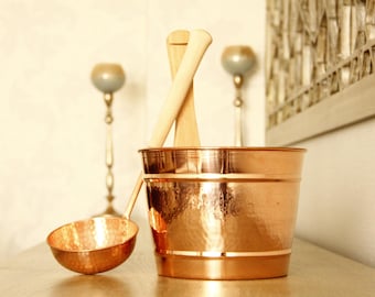 May include: A copper sauna bucket with a matching ladle and wooden handles. The bucket has a hammered texture and a shiny finish. Two wooden sauna spoons are placed inside the bucket. The sauna accessories are on a wooden surface.