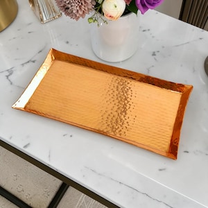 Könnte beinhalten: Ein rechteckiges Serviertablett mit kupferfarbenem Metallic-Finish und gehämmertem Muster. Das Tablett hat leicht erhöhte Ränder und eine reflektierende Oberfläche, die das Licht einfängt und ein markantes Muster auf dem Boden erzeugt.