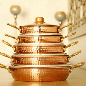 Peut inclure: Un ensemble de six pièces de cuisine en cuivre avec des surfaces martelées et des poignées dorées. Les ustensiles de cuisine sont empilés en forme de pyramide, la plus grande pièce en bas et la plus petite en haut.