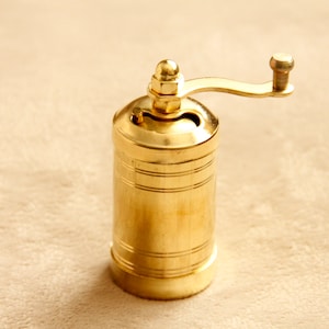 May include: A gold-colored manual pepper grinder with a crank handle. The grinder has a cylindrical shape and is made of metal.