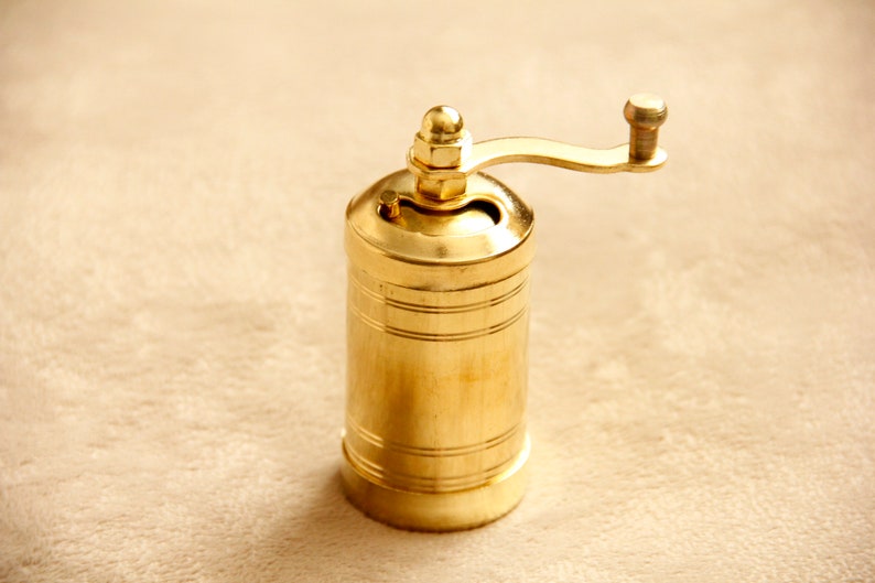 May include: A gold-colored manual pepper grinder with a crank handle. The grinder has a cylindrical shape and is made of metal.