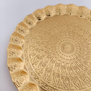 May include: A round, gold-colored metal serving tray with an intricate, embossed design. The tray has a scalloped edge and a raised center.