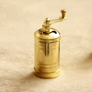May include: Two gold metal pepper grinders with a hand crank on top. The grinders are sitting on a beige surface.