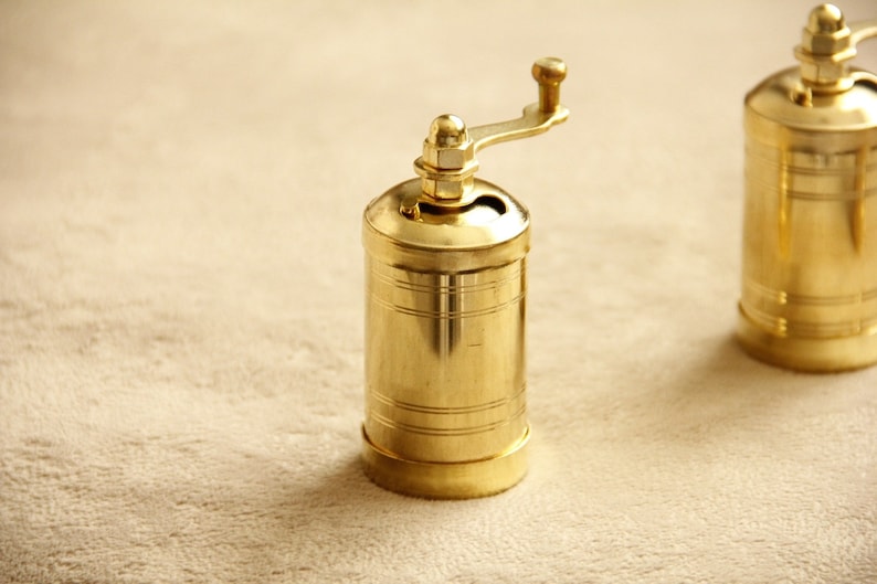 May include: Two gold metal pepper grinders with a hand crank on top. The grinders are sitting on a beige surface.
