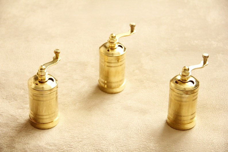 May include: Three gold-colored manual pepper grinders with a crank handle on a beige surface.