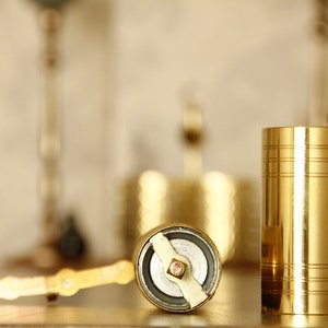 May include: A close-up of a gold-colored pepper grinder and a matching cylindrical container. The pepper grinder is open, revealing its grinding mechanism. The container has horizontal etched lines. The items are on a wooden surface.