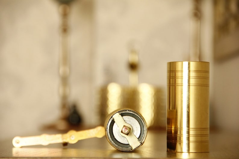 May include: A close-up of a gold-colored pepper grinder and a matching cylindrical container. The pepper grinder is open, revealing its grinding mechanism. The container has horizontal etched lines. The items are on a wooden surface.