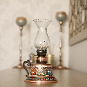 May include: A small, copper oil lamp with a painted floral design in shades of orange, blue, green, and black. The lamp has a clear glass chimney and a handle.
