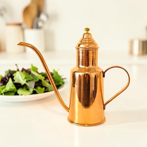 Puede incluir: Un dispensador de aceite de color cobre con un pico largo y curvo y un asa. El dispensador tiene una tapa con un pequeño pomo redondo en la parte superior. Un plato de ensalada está en el fondo. El dispensador está sobre una superficie blanca.