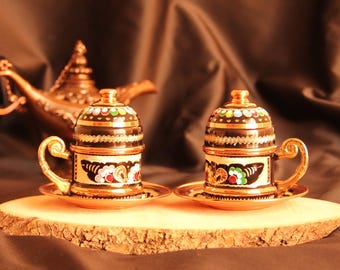 May include: Two ornate, gold-colored coffee cups with lids and saucers, featuring black and white floral designs. The cups sit on a wooden surface, with a decorative lamp in the background. The cups have a height of approximately 3 inches.