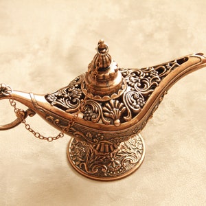 Peut inclure: Une lampe &agrave; genie de couleur dor&eacute;e avec des motifs floraux complexes et une cha&icirc;ne attach&eacute;e au bec. La lampe a une base ronde et un sommet pointu.