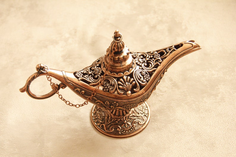 Peut inclure: Une lampe &agrave; genie de couleur dor&eacute;e avec des motifs floraux complexes et une cha&icirc;ne attach&eacute;e au bec. La lampe a une base ronde et un sommet pointu.