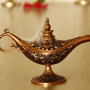 Peut inclure: Une lampe &agrave; genie dor&eacute;e avec des d&eacute;tails complexes et une poign&eacute;e en cha&icirc;ne. De la fum&eacute;e s'&eacute;l&egrave;ve du haut de la lampe.