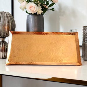 May include: A rectangular copper tray with a hammered texture and raised edges. The tray has a metallic finish with a warm orange-brown hue and a distinct dimpled surface pattern across its entire flat base and angled sides.