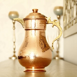May include: A hammered copper pitcher with a gold-toned handle and spout. The pitcher has a rounded body and a flared base.