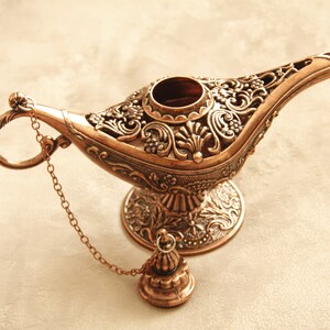 Peut inclure: Une lampe &agrave; genie de couleur dor&eacute;e avec des motifs floraux complexes et une cha&icirc;ne attach&eacute;e au bec. La lampe a une base ronde et un couvercle amovible.