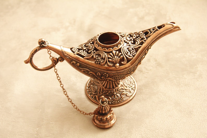 Peut inclure: Une lampe &agrave; genie de couleur dor&eacute;e avec des motifs floraux complexes et une cha&icirc;ne attach&eacute;e au bec. La lampe a une base ronde et un couvercle amovible.