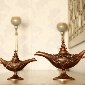 Peut inclure: Deux lampes &agrave; genie orn&eacute;es, de couleur or, avec des motifs complexes et une poign&eacute;e en cha&icirc;ne. Les lampes sont sur une surface brun clair.