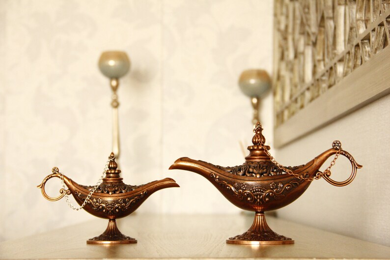 Peut inclure: Deux lampes &agrave; genie orn&eacute;es, de couleur or, avec des motifs complexes et une poign&eacute;e en cha&icirc;ne. Les lampes sont sur une surface brun clair.