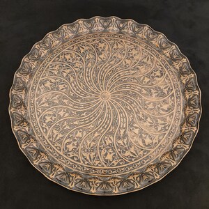 May include: A round, decorative tray with a black and gold design. The tray has a raised rim and a swirling pattern of leaves and flowers.