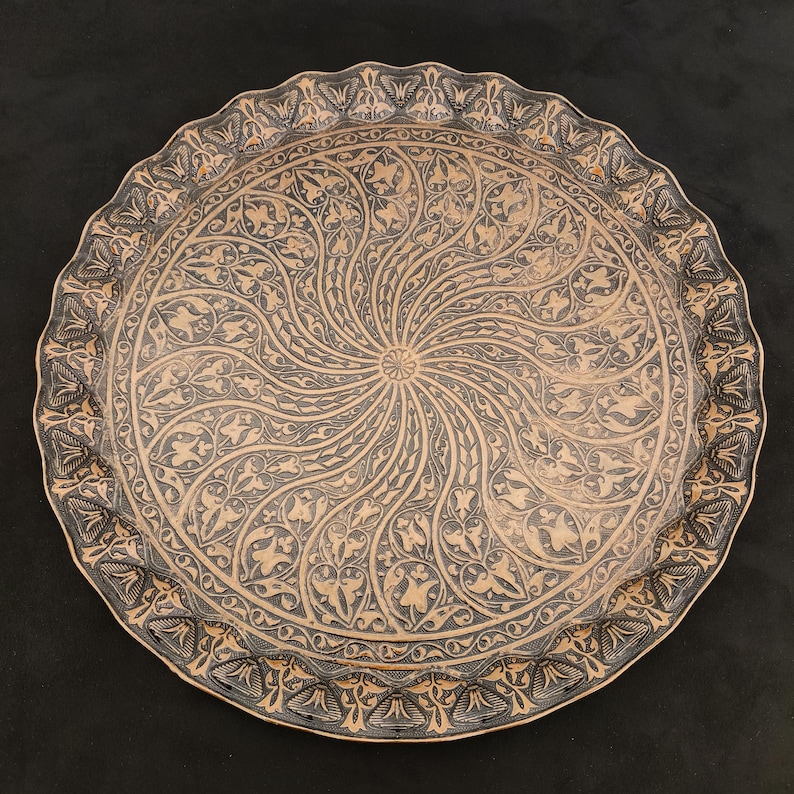 May include: A round, decorative tray with a black and gold design. The tray has a raised rim and a swirling pattern of leaves and flowers.
