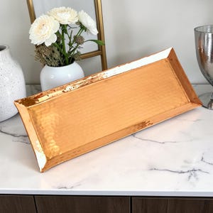 May include: A rectangular, hammered copper tray with a metallic finish and raised edges. The tray features a distinct textured surface created by the hammering process, reflecting light across its elongated, flat base and angled sides.