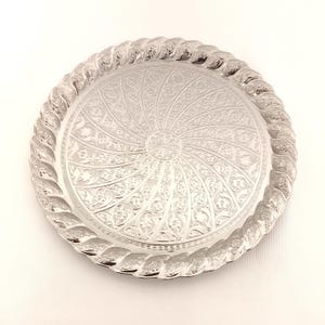 May include: A silver round serving tray with a decorative, intricate, swirling floral pattern embossed on the surface. The tray has a raised, rope-like border.