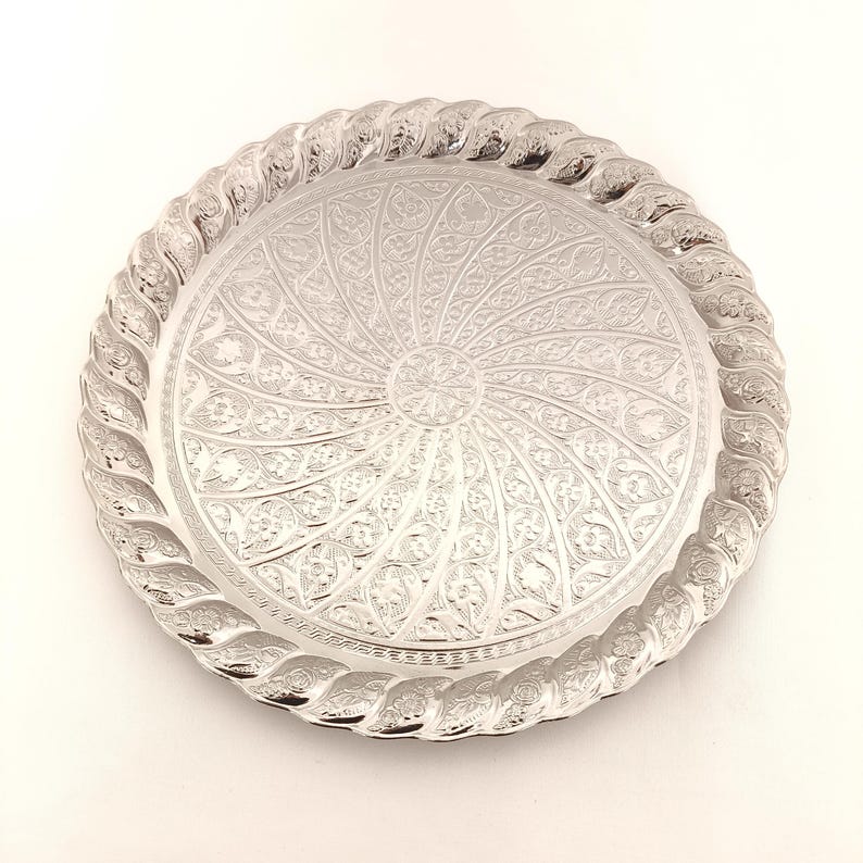 May include: A silver round serving tray with a decorative, intricate, swirling floral pattern embossed on the surface. The tray has a raised, rope-like border.