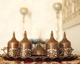 May include: Set of six ornate gold-toned metal coffee cups with lids, arranged on a tray with a beaded surface. The cups have intricate floral designs and are perfect for serving Turkish coffee.