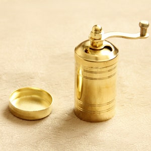 May include: A gold metal pepper grinder with a crank handle. The grinder has a cylindrical shape and is decorated with horizontal lines.